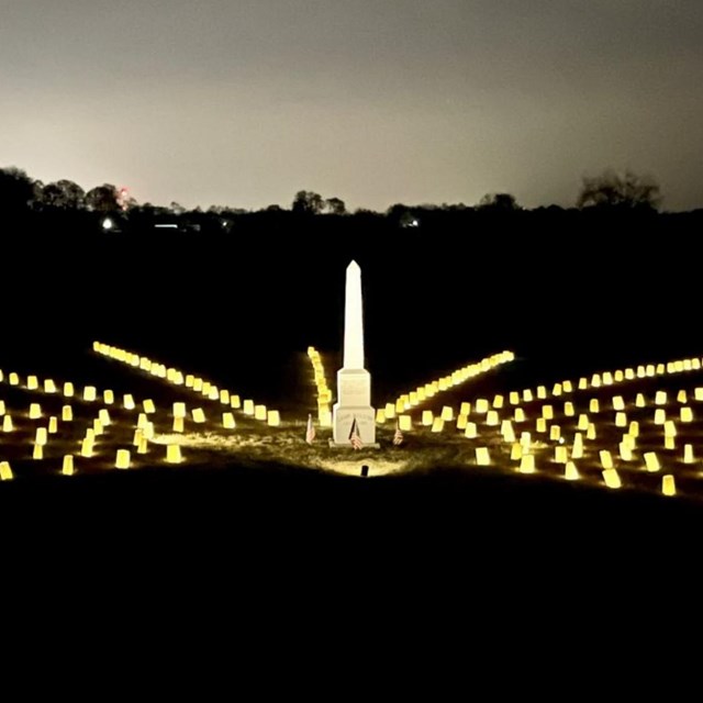 A stone obelisk surrounded by luminaries on a dark field with a bright light sky in the backgrond.