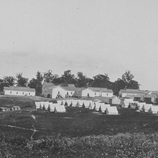 Wooden buildings and tents on a slight hill with tress in the background during the Civil War.