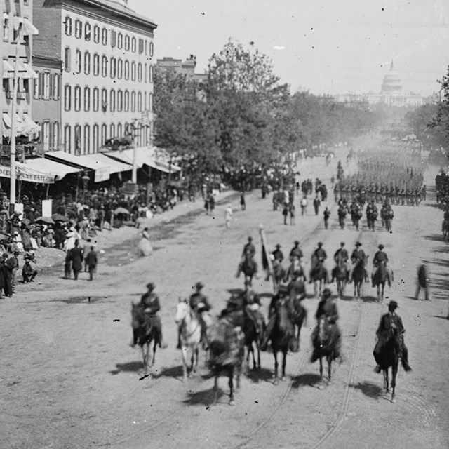 Soldiers mounted and on foot marching through a city street while civilians watch during Civil War.