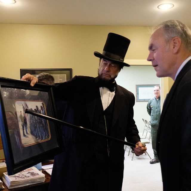 Man dressed as Abe Lincoln uses cane to point out framed artwork being observed by Ryan Zinke