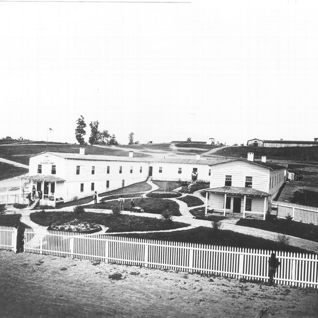 A U-shaped white wooden building with a lawn, foundation, and fence in front during the Civil War.