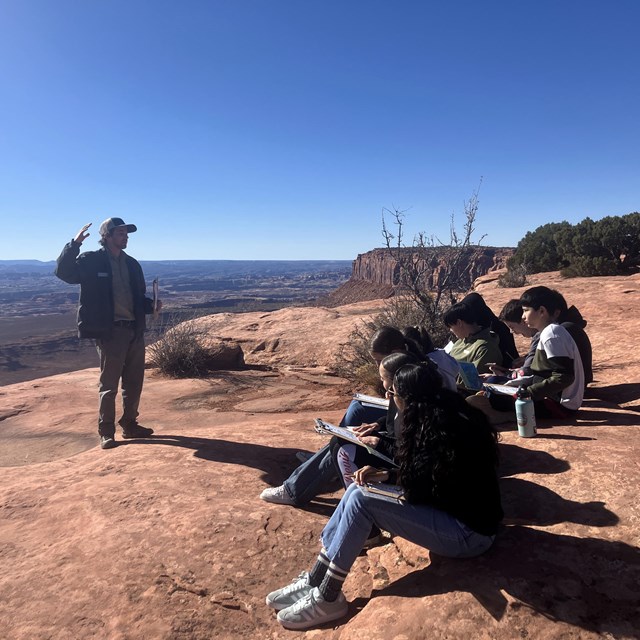 A person gives a presentation to a group of students sitting on brownish sandstone rocks.