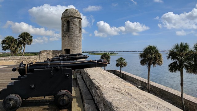 Image of gun deck with cannons, bell tower, water, and palm trees. 