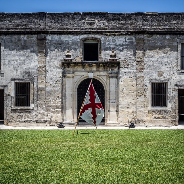 Grass in foreground, splanish flag centered in image, chapel door and walls in background