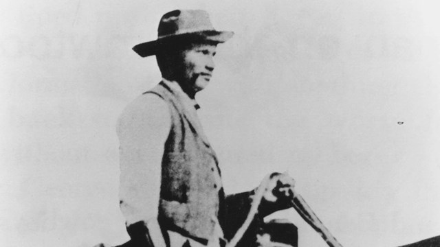 Black and white photo of cowboy with stirrups in hand on horse facing to the right. 