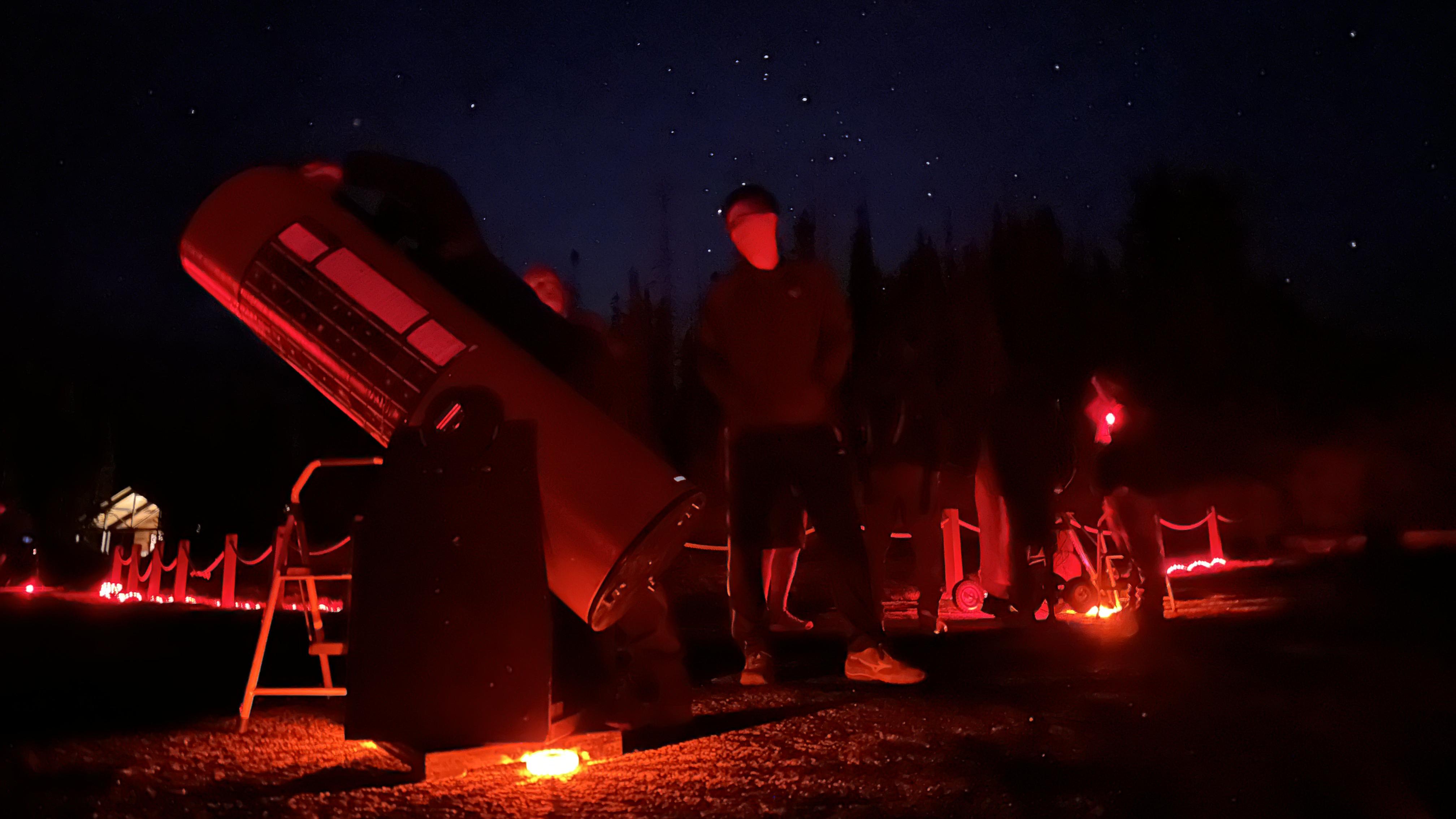 A person stands next to a telescope illuminated by red light before a starry dark sky.