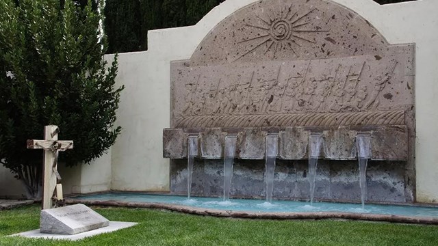 A gravestone with a cross sitting in green grass, next to a large water fountain.