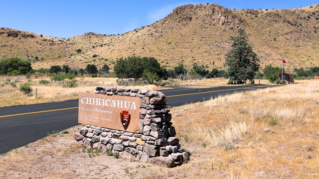 Brown and white sign in a rock structure, road, mountain and entrance booth behind