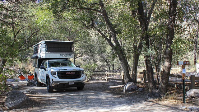 pop up camper on a white pick up truck with trees and 2 people sitting in chairs behind it