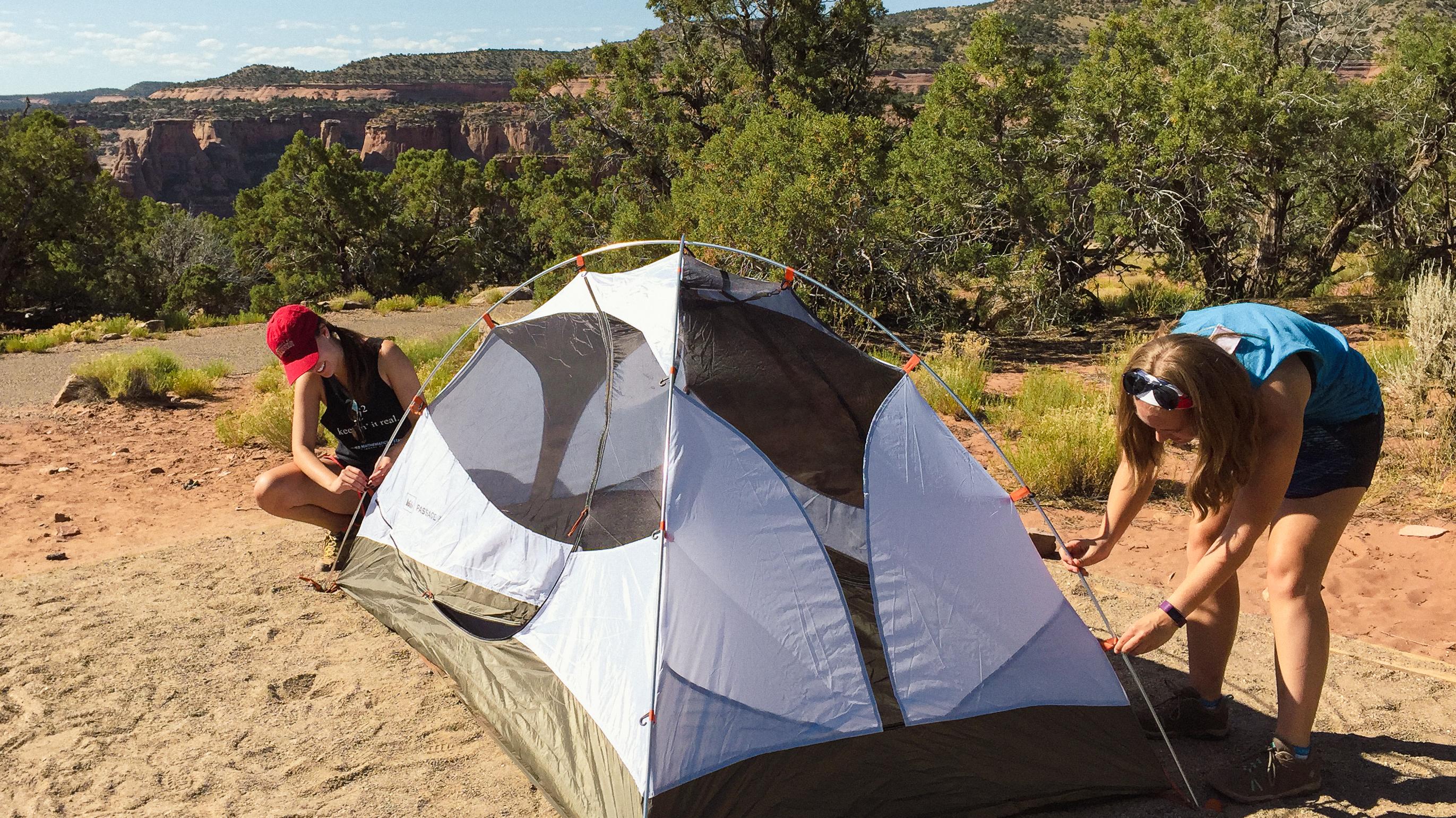 two people set up a small tent on a reddish dirt pad surrounded by green juniper trees