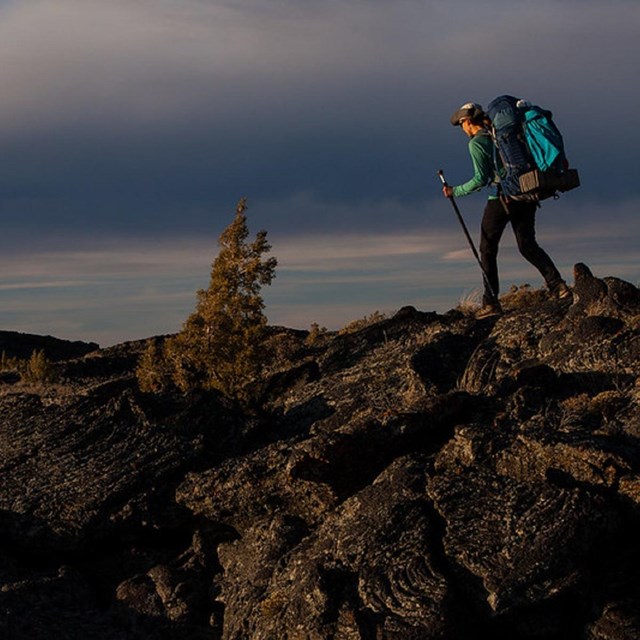a backpacker with a large backpack and walking stick traverses an expansive lava field @ golden hour