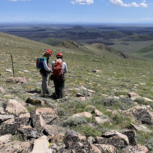 two people with backpacks wearing red helmets fixing a wire fence above a mountain vista