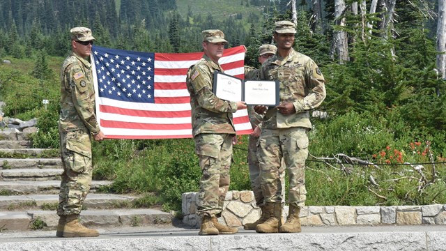 Soldiers holding a certificate and US flag in front of a mountain 