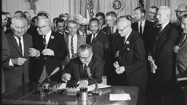 Historic photo of President Lyndon Johnson signing a bill with a crowd standing behind his desk