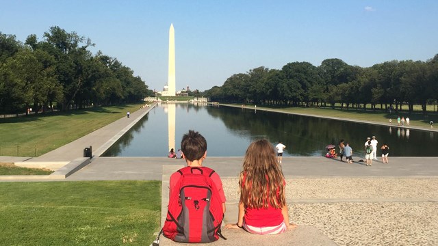 Two kids sitting on a memorial step with the Washington Monument in the distance