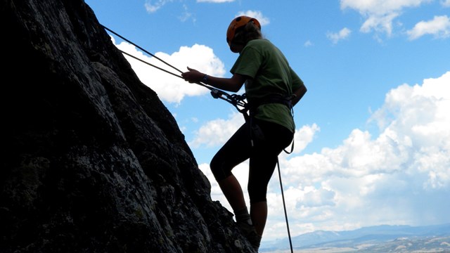 A solo rock climber wearing an orange helmet using ropes to descend the side of a mountain.