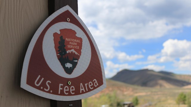 A 'U.S. Fee Area' triangular metal sign with an arrowhead logo. Sign is mounted to a wall.