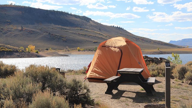 An orange tent is pitched behind a picnic table and fire grate. Sagebrush and water are behind it.