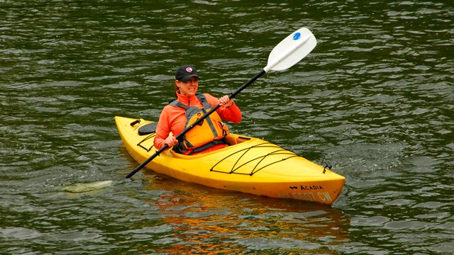 A person in a bright yellow kayak in the water, holding a paddle and wearing an orange life vest.