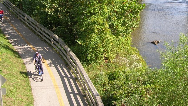 A person shown from above, riding a bicycle on a paved trail next to a tree-lined river.