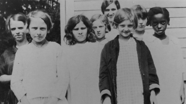 Seven girls in plain dresses crowd in front a woman who is only a head taller.