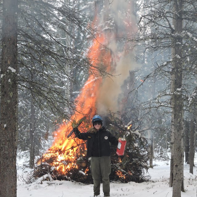 A woman in grey pants and a black shirt and jacket stands in front of a burning pile of brush.