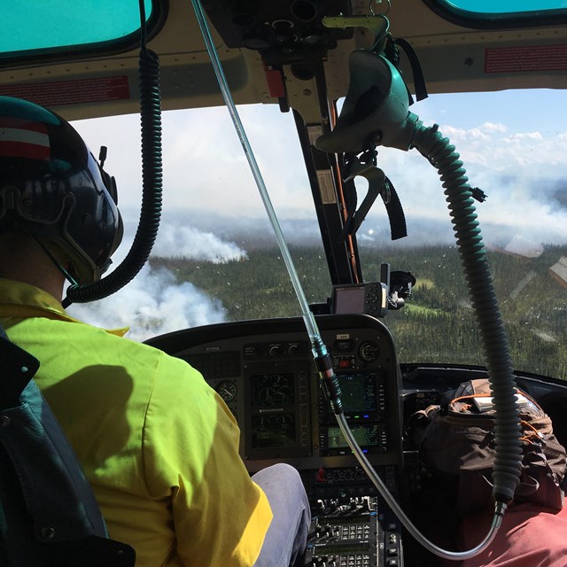 A helicopter pilot in a neon yellow shirt looks out a window towards smoke rising from the trees.