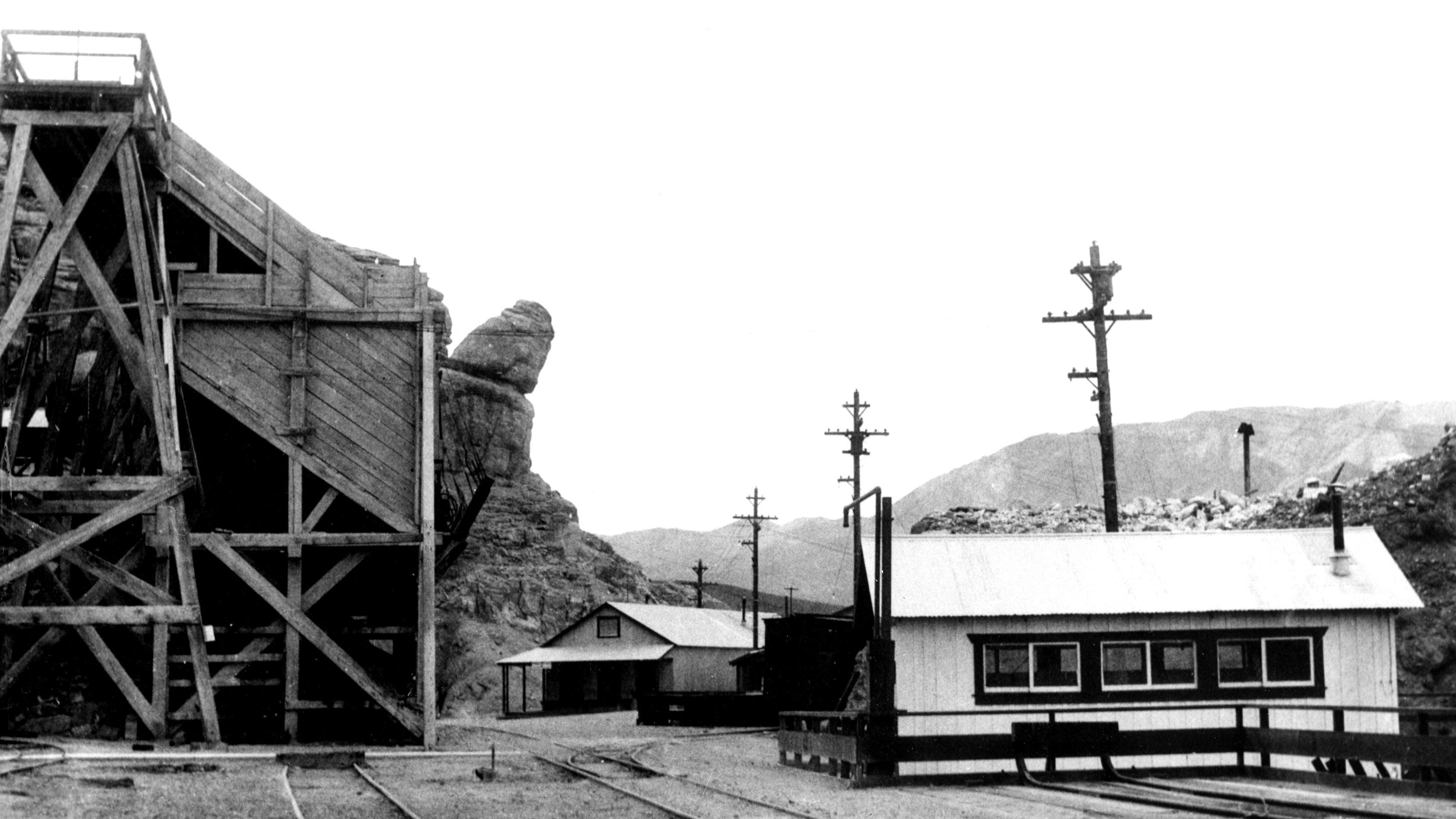 A black-and-white photograph depicts a small, rural railway station set against a barren hillside. 
