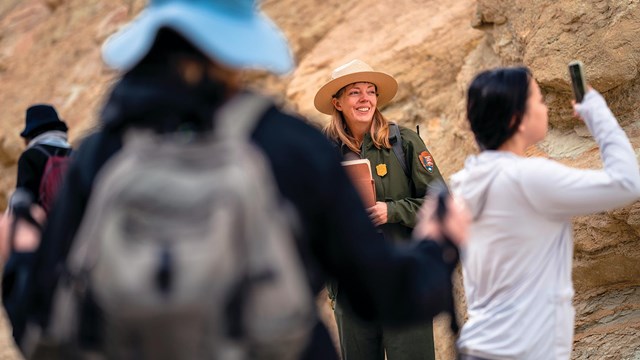 Uniformed ranger smiles while giving a tour in a yellow canyon to visitors wearing hiking gear.