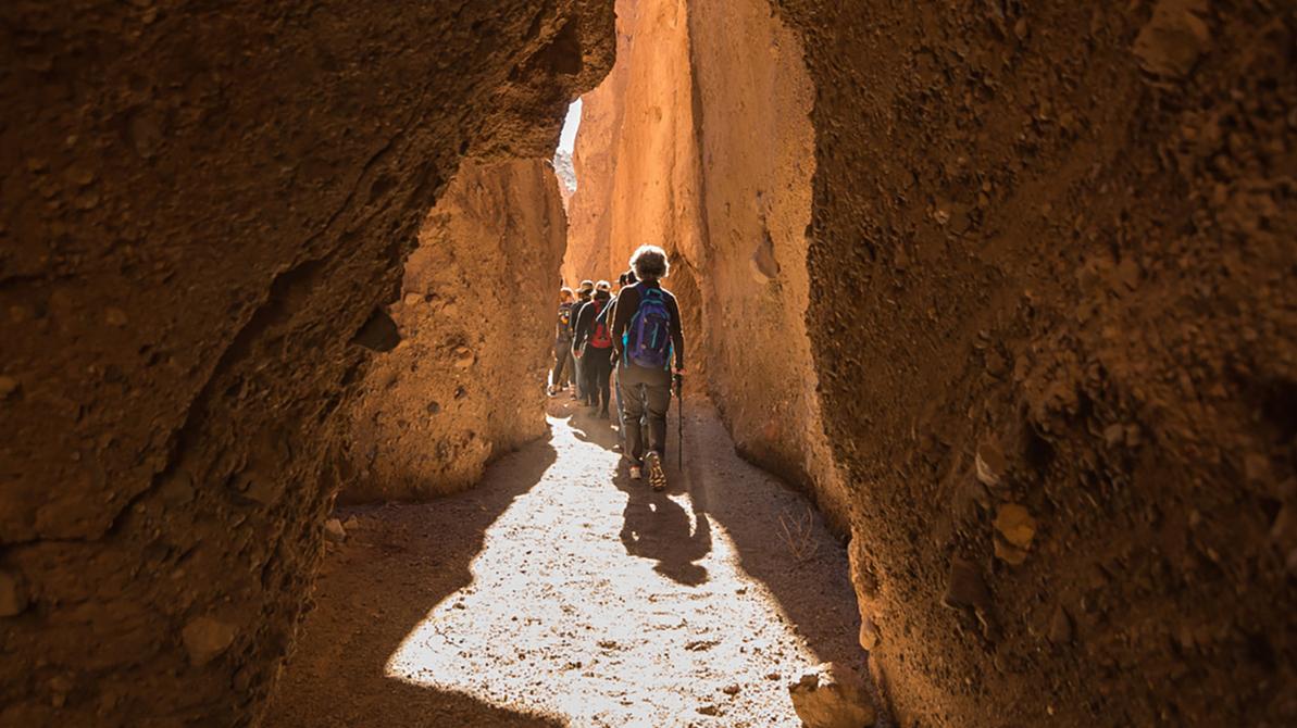 Seen from deep shade, a single-file line of hikers moves away through a tight rock passage. 