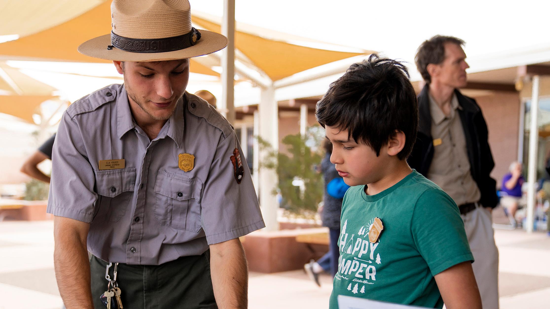 A young park ranger leans over an outreach table and points to a handout as a boy listens beside him