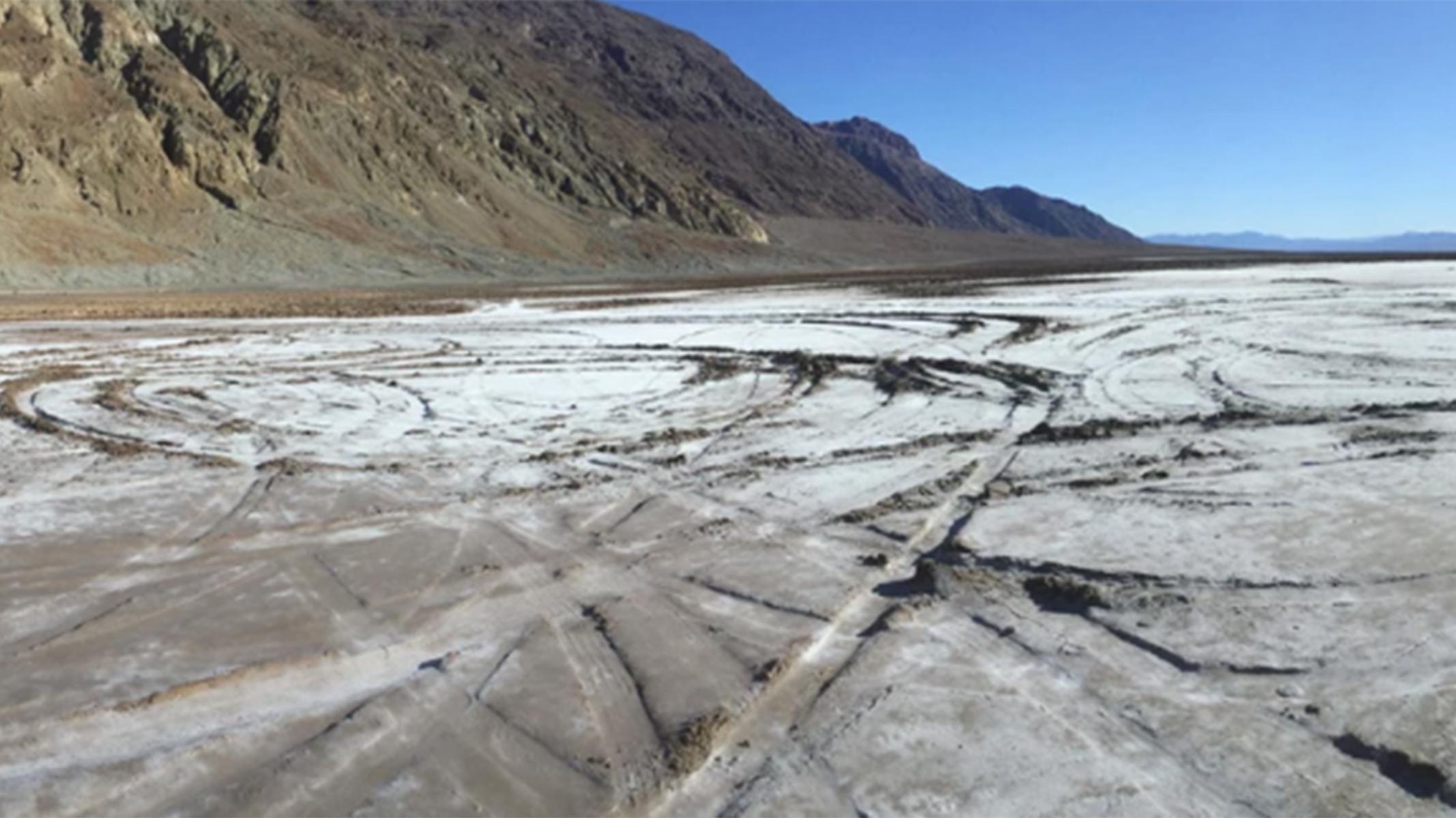  A vast, pale, crusted flat fills most of the frame, etched with looping vehicle tracks. 