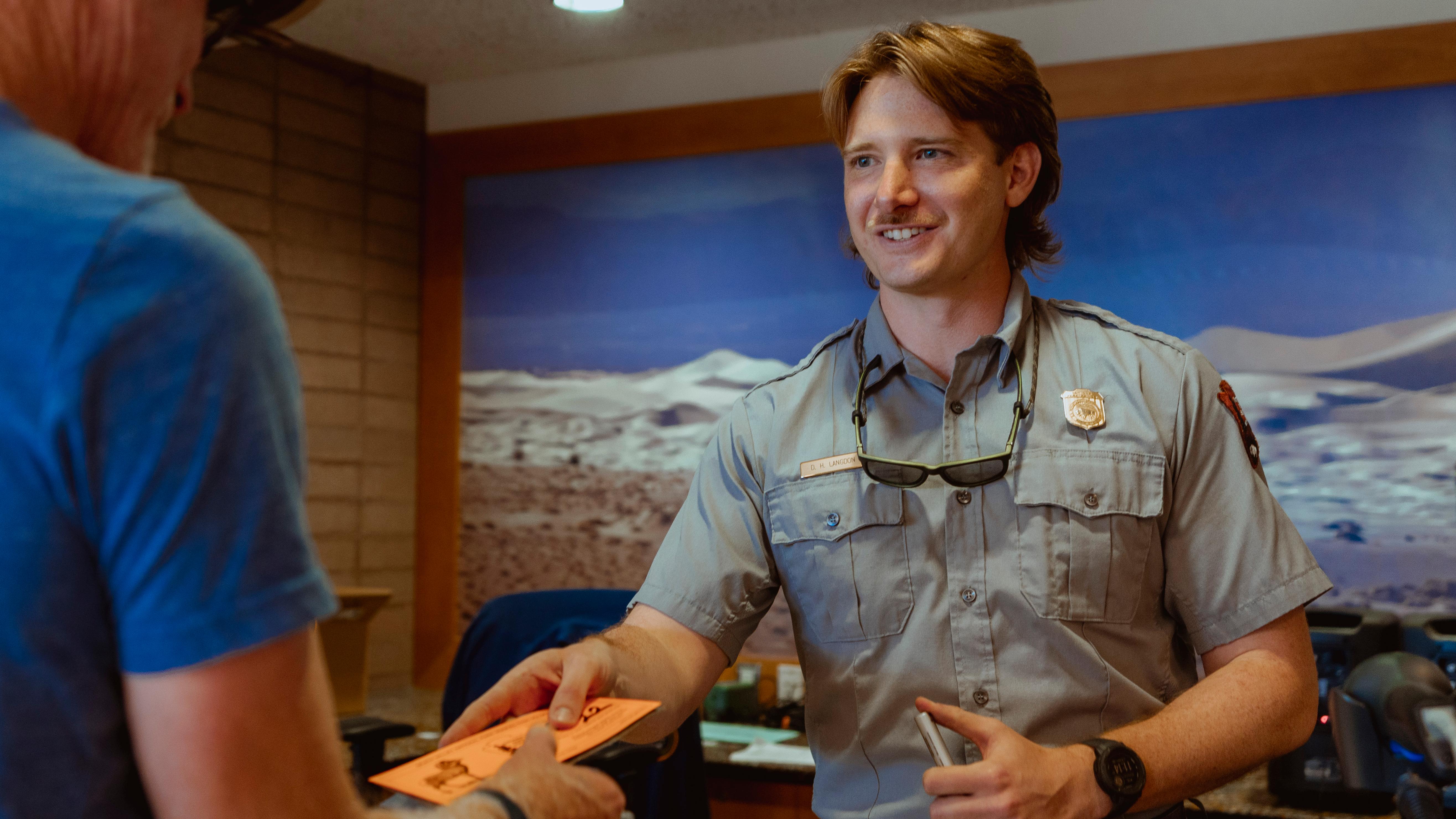 A smiling park ranger in uniform hands a paper pass or ticket to a visitor standing at a counter.