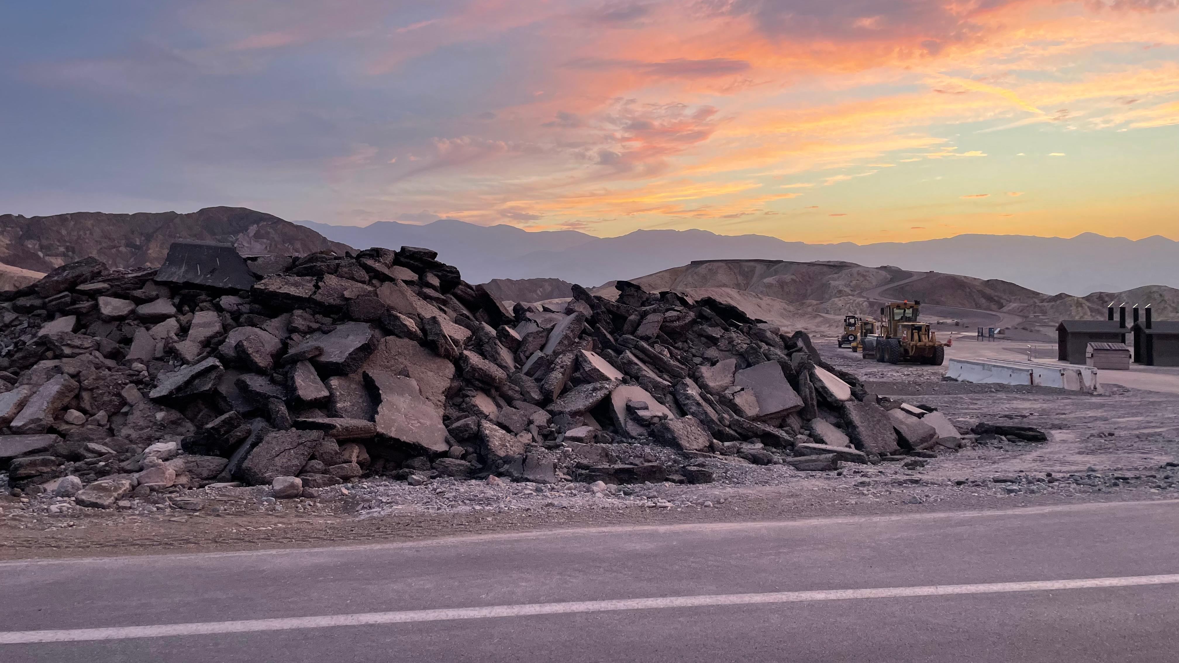 Pile of broken pavement along the side of a road under a pink, yellow and blue sunset.