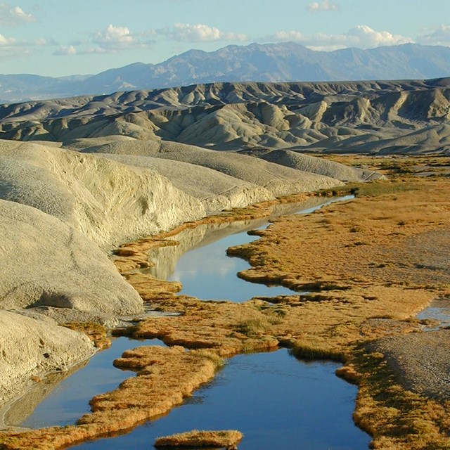 A deep blue creek with brown vegetation winds through the tan hills of a desert with mountains.