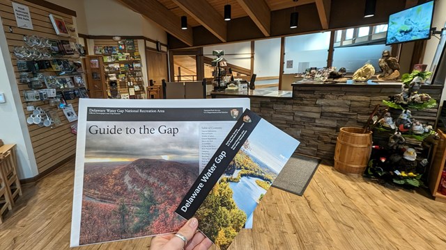 A hand holding out a guide and a park map with the visitor center in the background.