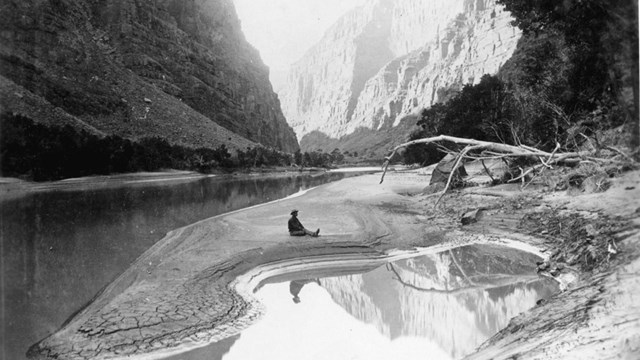 A black and white photograph of Lodore Canyon with a man sitting on a sandbar in the river. 