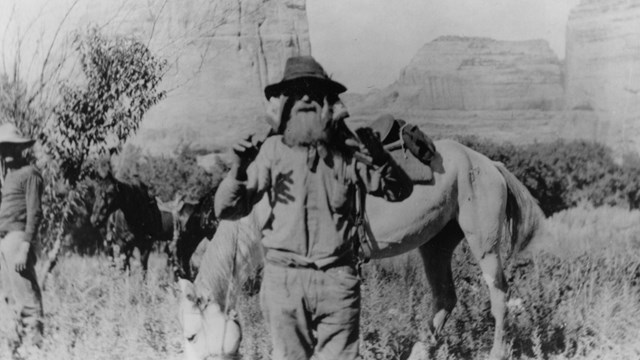 A black and white photo of a man with a grizzled beard in front of a horse.