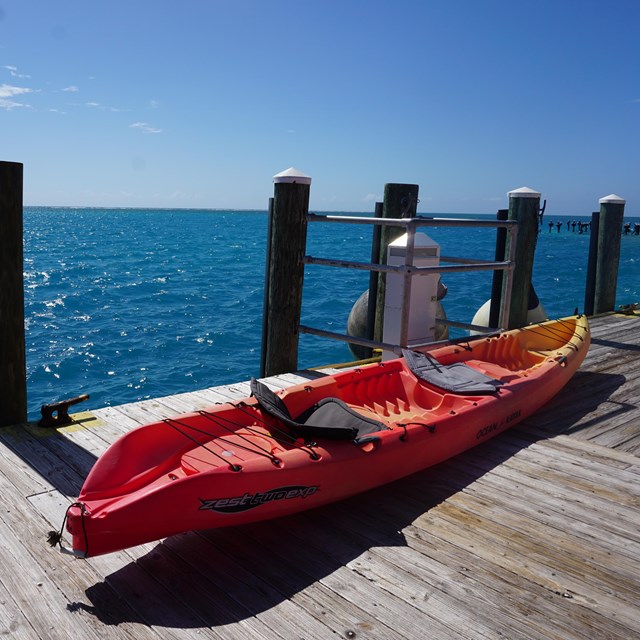 A long orange and yellow kayak vessel resting on a wooden dock beside a blue ocean and blue sky
