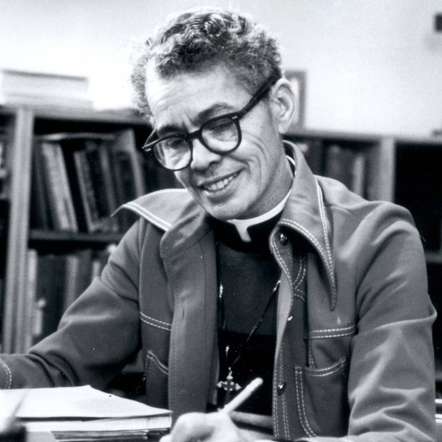 A portrait photograph of Pauli Murray seated at a desk with pen in her hand.