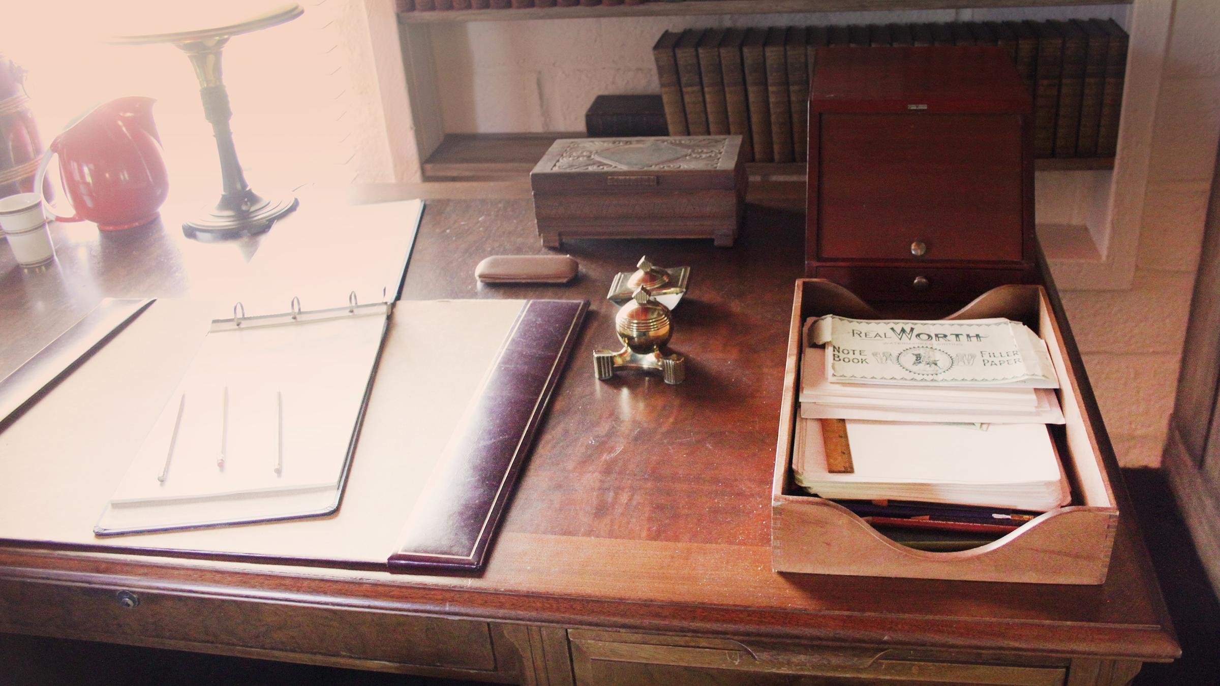 Wooden desk with organized papers, a lamp, and vintage office supplies, surrounded by bookshelves.