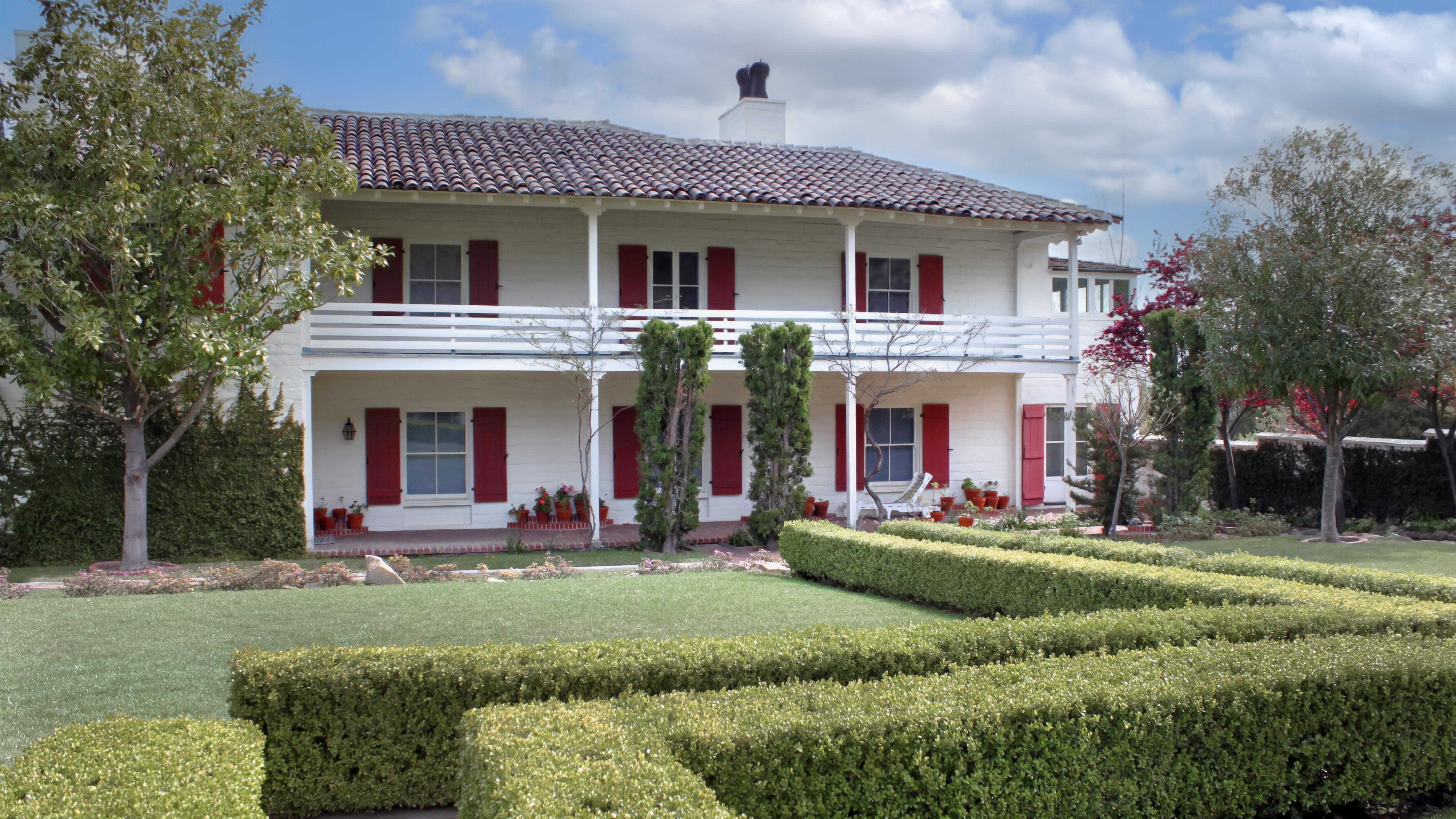 Two-story white house with red shutters, a tile roof, and a landscaped garden in front.
