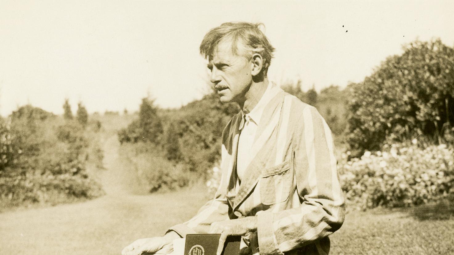 Vintage photo of a man in a striped jacket holding a book, standing in a garden with trees.