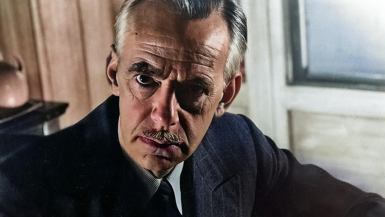 Portrait of a serious man with a mustache, wearing a suit and tie, seated indoors near a window.