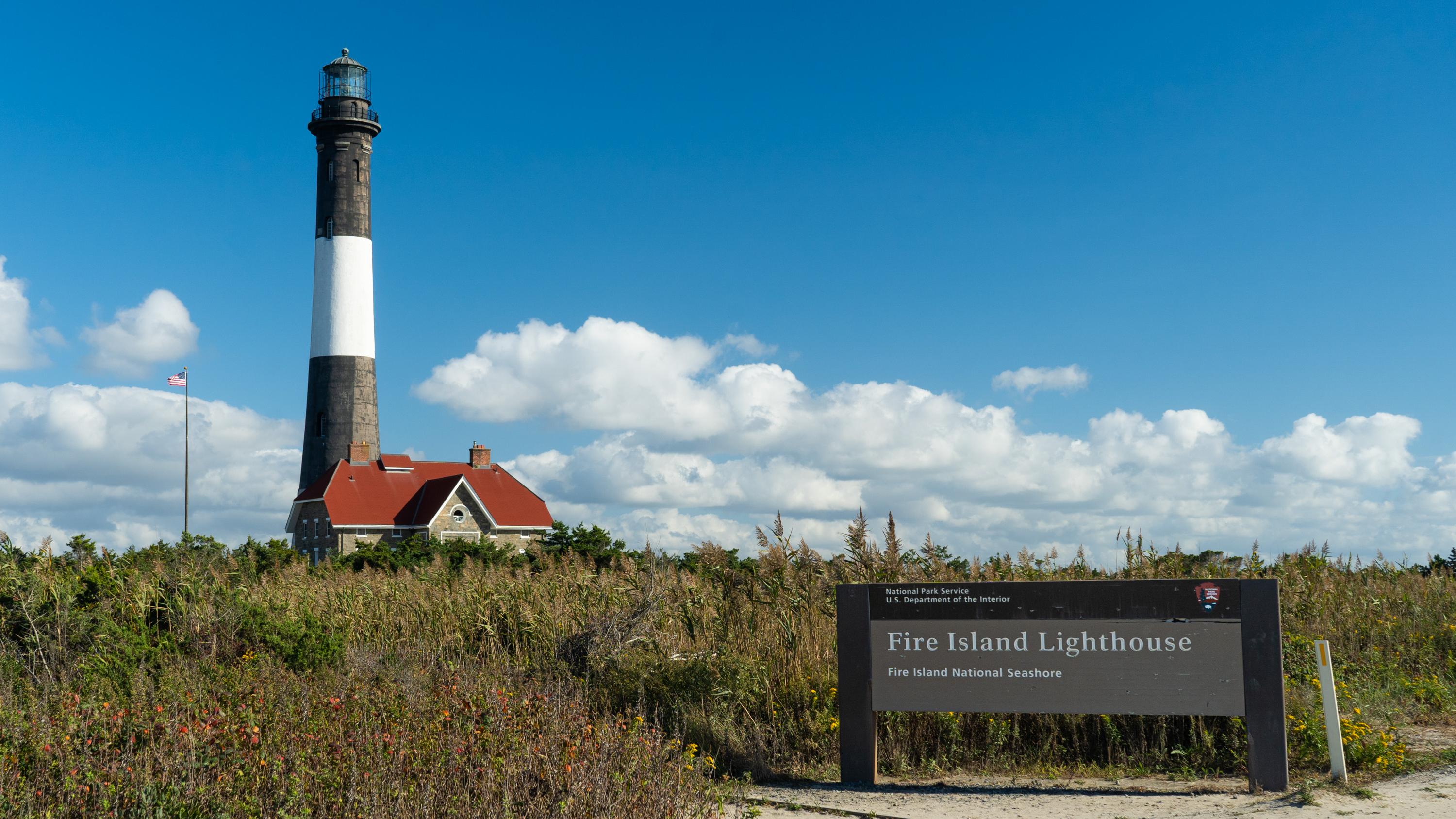 A sign reading "Fire Island Lighthouse" with a black and white striped lighthouse in the background.