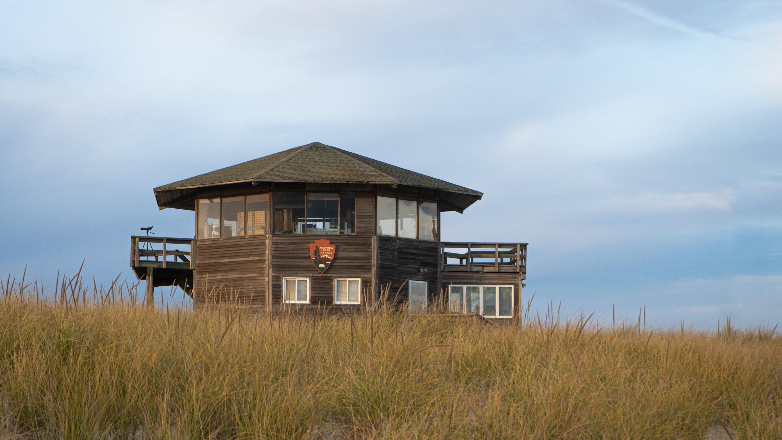 A wooden building peeks out from behind a grass-covered dune with blue sky above.