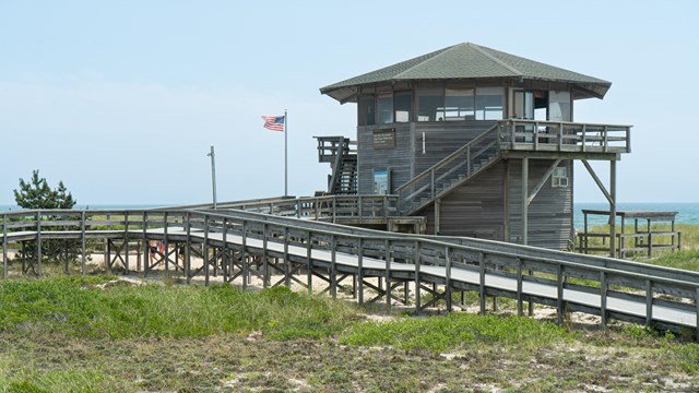 An octagonal shaped, 2-story wooden building with sand dunes and broadwalk in view.