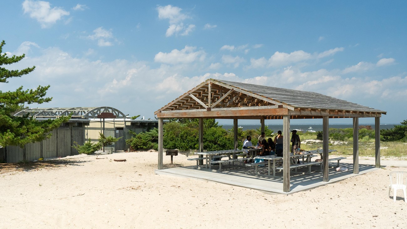 A open shade structure with picnic tables in a sandy area with blue sky and clouds in the background