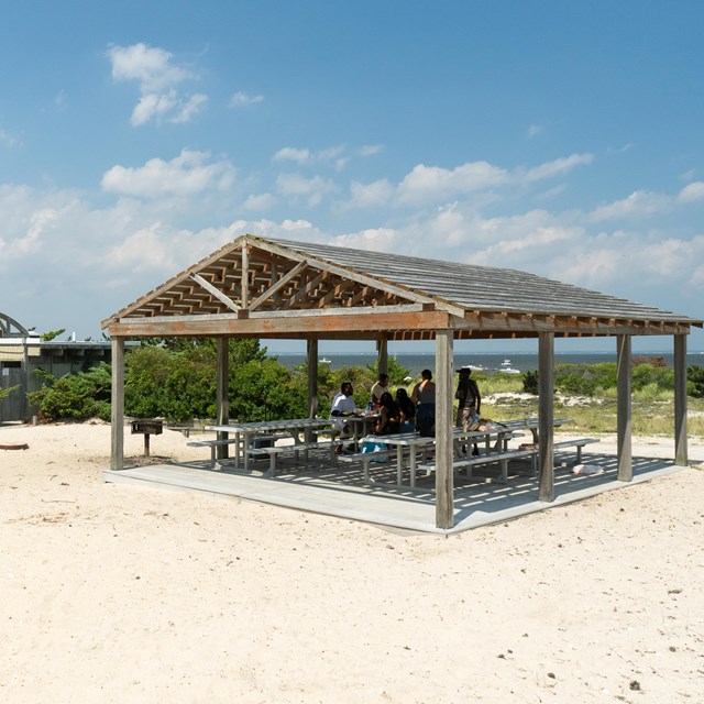 A open shade structure with picnic tables in a sandy area with blue sky and clouds in the background