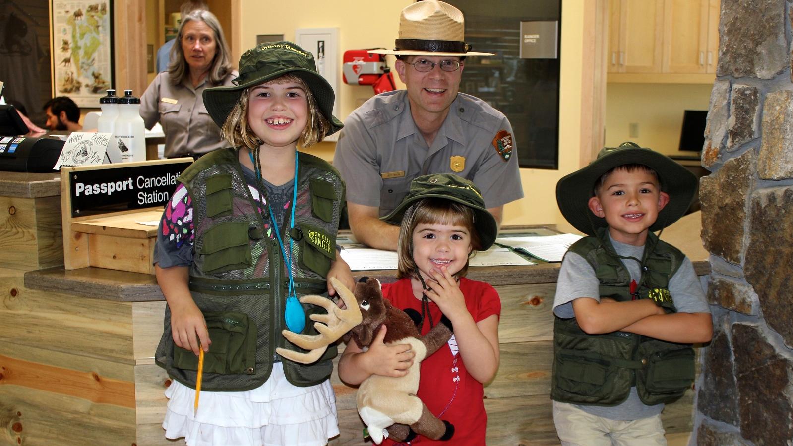 Three children wearing hats in front of a desk with a male and female park ranger behind.